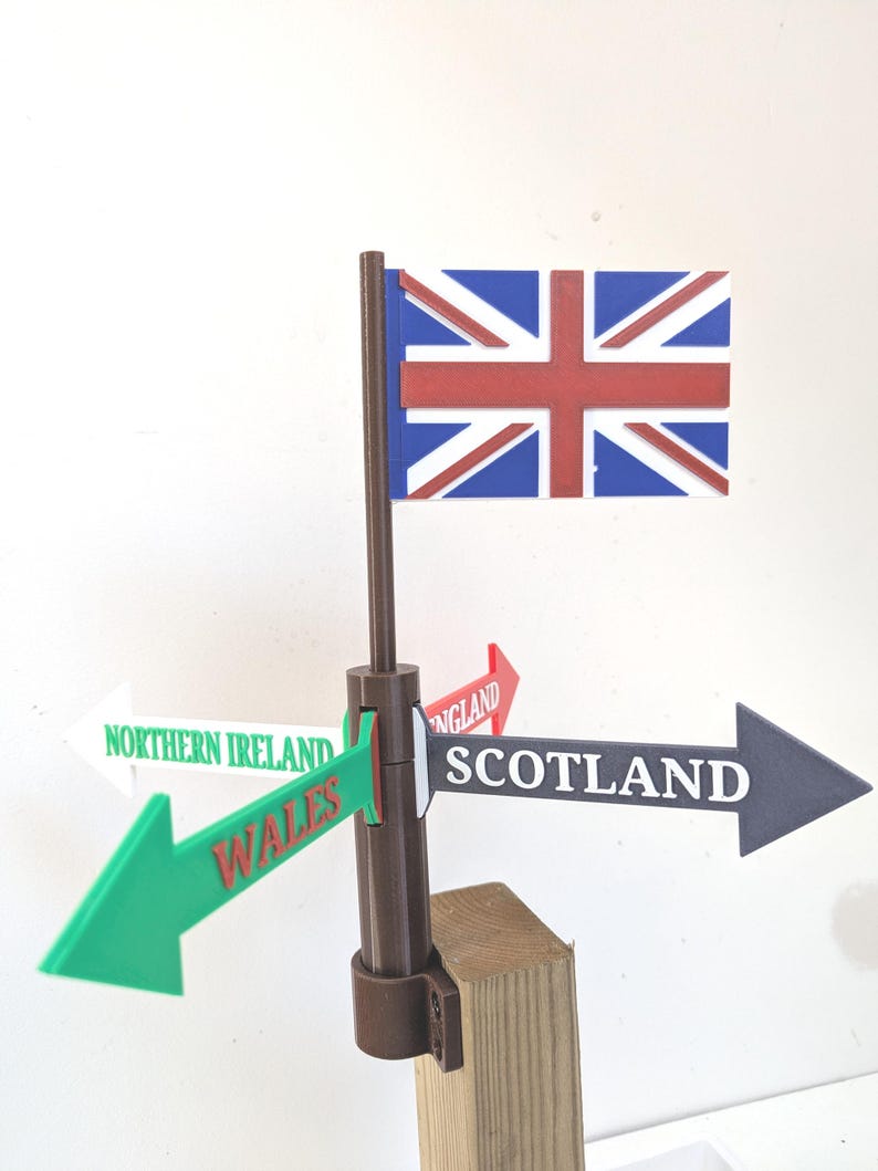 May include: A brown plastic signpost featuring a Union Jack flag at the top and four directional arrows pointing outward. The arrows are labelled with the text Northern Ireland, Wales, England, and Scotland in various colours and fonts.