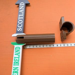 May include: Two directional arrow signs labelled SCOTLAND and NORTHERN IRELAND, a brown cylindrical connector, and a brown mounting bracket. A white measuring tape shows the connector is approximately 24 cm long, with markings up to 16 cm.