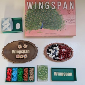 Op de afbeelding: Het bordspel "Wingspan Asia" met een roze doos met een pauwenillustratie. Bevat spelstukken, kaarten en bakjes in verschillende kleuren. De spelcomponenten zijn gerangschikt op een wit oppervlak.