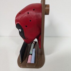 May include: A red and black Deadpool-themed chalk holder. The holder features a red head with black eye cutouts and a brown base. It contains several pieces of chalk in various colors, including pink, blue, and green.