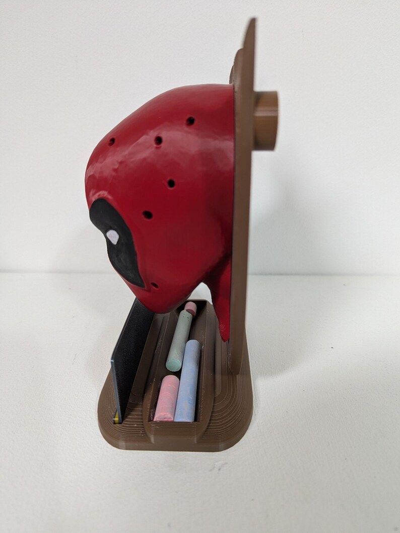 May include: A red and black Deadpool-themed chalk holder. The holder features a red head with black eye cutouts and a brown base. It contains several pieces of chalk in various colors, including pink, blue, and green.