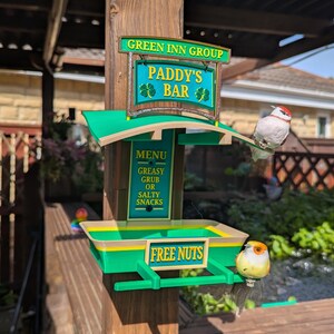 Peut inclure: Une mangeoire à oiseaux verte et beige en forme d'enseigne de pub avec le texte GREEN INN GROUP, PADDY'S BAR, MENU GREASY GRUB OR SALTY SNACKS, et FREE NUTS. Deux petits oiseaux artificiels sont posés sur la structure fixée à un poteau en bois.