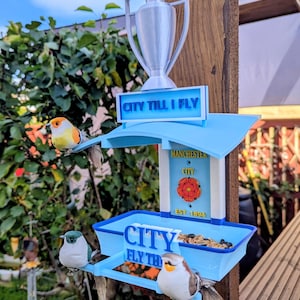 Op de afbeelding: Lichtblauwe vogelvoederhuisje met de tekst "CITY" en "CITY TILL I FLY". De voederbak heeft een trofee en een bord met "Manchester City". Decoratieve vogels zitten op de voederbak. De voederbak is op een houten paal gemonteerd.