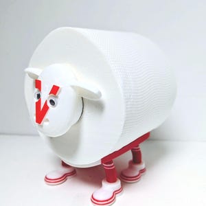 Può includere: Un portarotolo di carta igienica a forma di pecora bianca. La pecora ha una 'V' rossa sul viso, occhi mobili, gambe rosse e scarpe bianche con strisce rosse. Il rotolo di carta igienica forma il corpo della pecora.