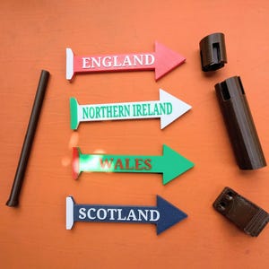 May include: A set of four directional arrow signs labelled England, Northern Ireland, Wales, and Scotland, accompanied by a long dark brown rod, a cylindrical connector, and a rectangular base piece, all arranged on a flat orange surface.