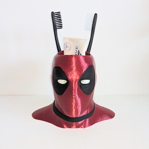 May include: A 3D printed Deadpool head, in red and black, against a white background. The head is used as a toothbrush holder and contains two toothbrushes and a tube of toothpaste.