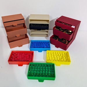 May include: A collection of board game accessories, including small storage boxes in shades of brown, beige, and red. Also, there are trays filled with blue, red, yellow, and green game pieces. The pieces appear to be small cubes.