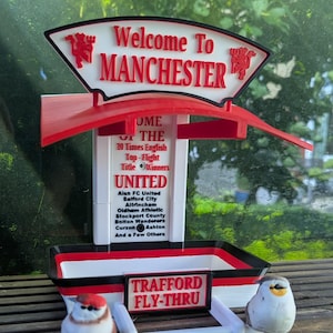 May include: A white, red, and black bird feeder featuring a sign that reads Welcome To MANCHESTER with two red devil emblems. The central pillar displays text about English football title winners and local clubs, with a sign reading TRAFFORD FLY-THRU.