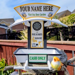 Peut inclure: Un panneau en plastique blanc et noir avec une arche supérieure portant le texte YOUR NAME HERE et Visit Our Beer Garden. Le panneau central affiche un graphique de cible avec POOL & DARTS, KARAOKE et LIVE MUSIC. Un panneau indique CASH ONLY.