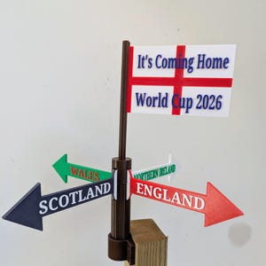 May include: A brown plastic signpost with four directional arrows pointing to Scotland, Wales, Northern Ireland, and England. At the top, a white flag with a red cross features the blue text It's Coming Home and World Cup 2026.