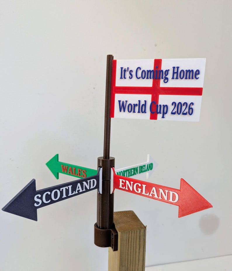 May include: A brown plastic signpost with four directional arrows pointing to Scotland, Wales, Northern Ireland, and England. At the top, a white flag with a red cross features the blue text It's Coming Home and World Cup 2026.