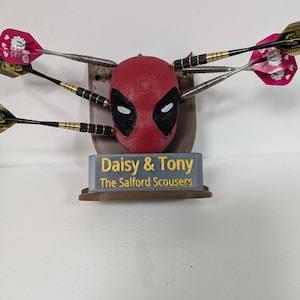 May include: A red Deadpool dartboard with black eye and mouth details, surrounded by darts. The base has the text "Daisy & Tony The Salford Scousers". The darts have pink and green flights.