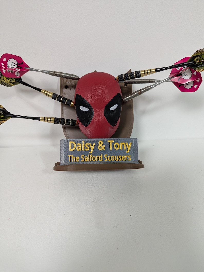 May include: A red Deadpool dartboard with black eye and mouth details, surrounded by darts. The base has the text "Daisy & Tony The Salford Scousers". The darts have pink and green flights.