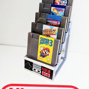 Op de afbeelding: Een grijze NES-game cartridgehouder met een witte basis met het NES-controllerontwerp. De houder toont verschillende klassieke game cartridges, waaronder Super Mario Bros. 3 en Gremlins 2. Onderaan staat een rood Nintendo-logo.