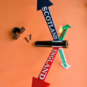 May include: A directional signpost featuring four coloured arrows pointing in different directions. The arrows are labelled SCOTLAND in blue, WALES in green, ENGLAND in red, and NORTHERN IRELAND in green. Includes a brown mounting bracket and two screws.
