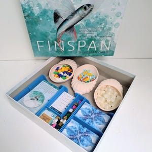 以下が含まれることがあります： ボードゲーム「Wingspan」が表示され、ターコイズと白のデザインとゲームタイトルが特徴の箱があります。箱の中には、カード、トークン、カラフルなピースが入った容器など、さまざまなゲームコンポーネントがあります。ゲームのテーマは鳥です。