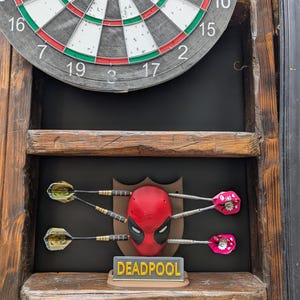 May include: A dartboard with a wooden frame, featuring a Deadpool-themed dart holder. The holder displays a red Deadpool mask with darts and a sign that reads "DEADPOOL". The darts have gold and pink flights.