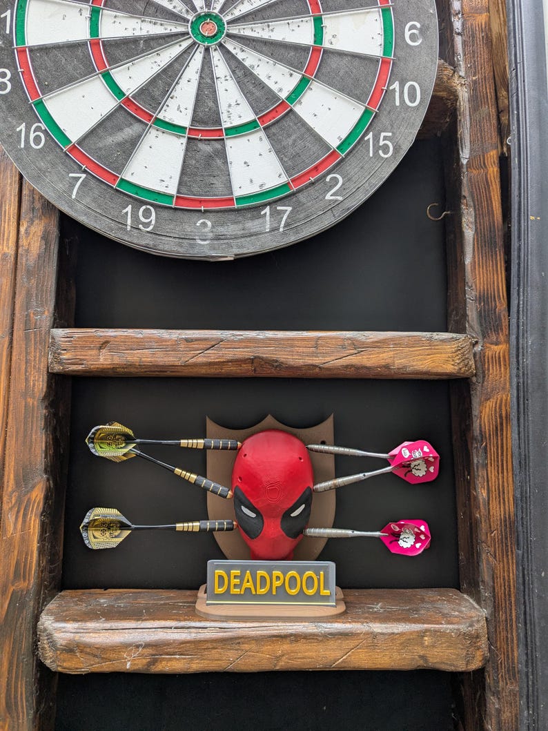 May include: A dartboard with a wooden frame, featuring a Deadpool-themed dart holder. The holder displays a red Deadpool mask with darts and a sign that reads "DEADPOOL". The darts have gold and pink flights.
