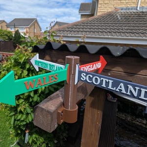 May include: A multi-directional signpost featuring four arrow-shaped signs pointing in different directions. The signs are labelled Wales in orange, Northern Ireland in green, England in red, and Scotland in dark blue, all mounted on a brown post.