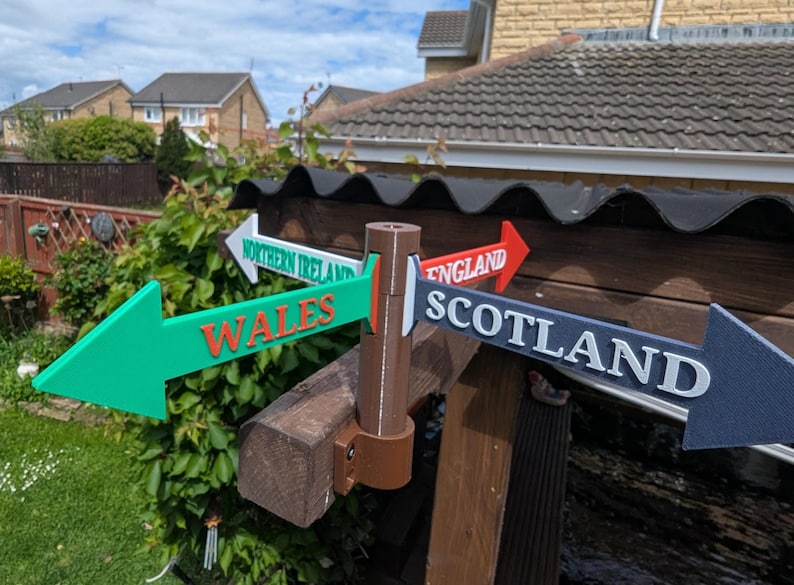 May include: A multi-directional signpost featuring four arrow-shaped signs pointing in different directions. The signs are labelled Wales in orange, Northern Ireland in green, England in red, and Scotland in dark blue, all mounted on a brown post.