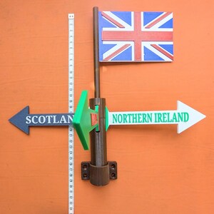 May include: A brown plastic signpost featuring a Union Jack flag at the top and three directional arrows pointing to Scotland, Wales, and Northern Ireland. A white measuring tape is placed vertically next to the post, showing a height of 34 cm.