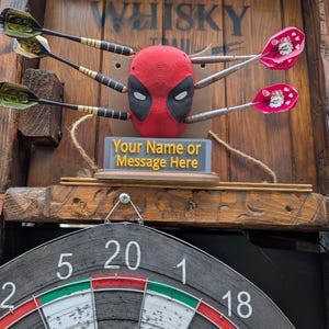 May include: A dartboard set with a Deadpool-themed target. The target is red with black eye cutouts, and the darts are embedded in it. The dartboard is visible below, with the text "Your Name or Message Here". The background is wooden.
