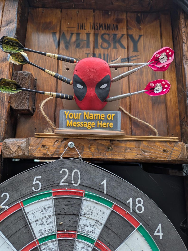 May include: A dartboard set with a Deadpool-themed target. The target is red with black eye cutouts, and the darts are embedded in it. The dartboard is visible below, with the text "Your Name or Message Here". The background is wooden.