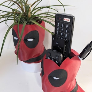 May include: Red and black Deadpool-themed planter and remote control holder. The planter holds a green plant with long leaves. The remote control holder has a pen and two remotes inside. The items are on a white surface.