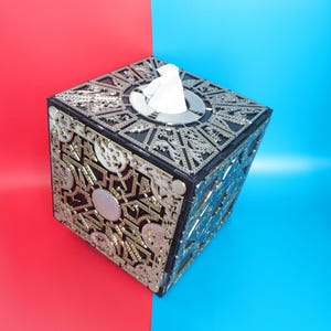 May include: A black and silver tissue box shaped like a puzzle cube with a white tissue peeking out of the top opening.