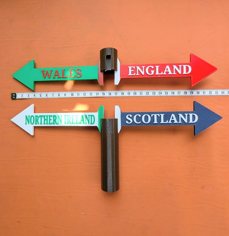 May include: Two directional sign sets with brown central posts. The top set features green and red arrows labelled WALES and ENGLAND. The bottom set features white and blue arrows labelled NORTHERN IRELAND and SCOTLAND. A measuring tape shows a width of 34 cm.