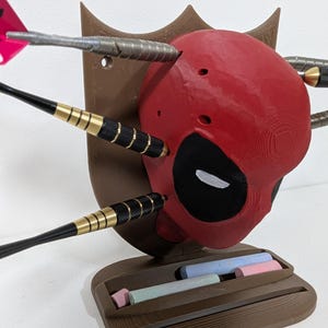 May include: A red Deadpool head with black eye cutouts and darts embedded in it. The head is mounted on a brown plaque with a small shelf holding colorful chalk sticks. The darts have black and gold accents.