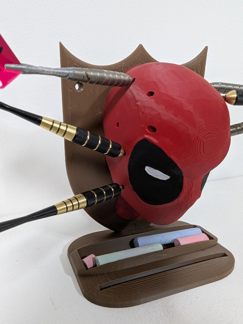 May include: A red Deadpool head with black eye cutouts and darts embedded in it. The head is mounted on a brown plaque with a small shelf holding colorful chalk sticks. The darts have black and gold accents.