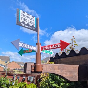 May include: A brown post with a rectangular sign at the top reading HOWAY THE LADS NEWCASTLE. Below are four directional arrows pointing in different directions labelled SCOTLAND, ENGLAND, WALES, and NORTHERN IRELAND in various colours.