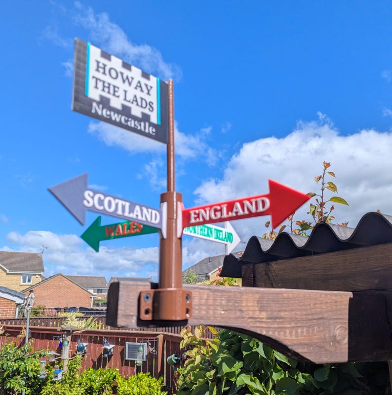 May include: A brown post with a rectangular sign at the top reading HOWAY THE LADS NEWCASTLE. Below are four directional arrows pointing in different directions labelled SCOTLAND, ENGLAND, WALES, and NORTHERN IRELAND in various colours.