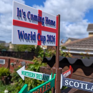 May include: A brown plastic signpost featuring a white rectangular flag with a red cross and the blue text It's Coming Home World Cup 2026. Below the flag are directional arrows labelled Northern Ireland, Wales, Scotland, and England.