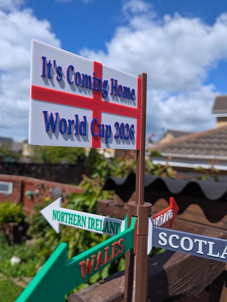 May include: A brown plastic signpost featuring a white rectangular flag with a red cross and the blue text It's Coming Home World Cup 2026. Below the flag are directional arrows labelled Northern Ireland, Wales, Scotland, and England.