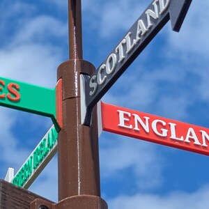 May include: A brown vertical post featuring multiple directional arrow signs pointing in different directions. The signs are coloured red, green, and grey, with white text reading Scotland, England, and Wales clearly visible on the directional markers.