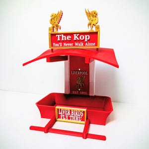 May include: A red and gold bird feeder featuring the text "The Kop" and "You'll Never Walk Alone." The feeder also displays the text "Liver Birds Fly Thru" and the Liverpool crest. The feeder is made of plastic and is a fan item.
