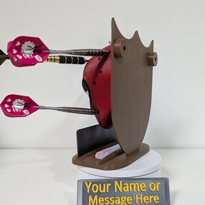 May include: A red and black Deadpool-themed dart holder with three darts. The holder has a brown shield-like back and a base with a slot for darts. A sign at the base reads "Your Name or Message Here."