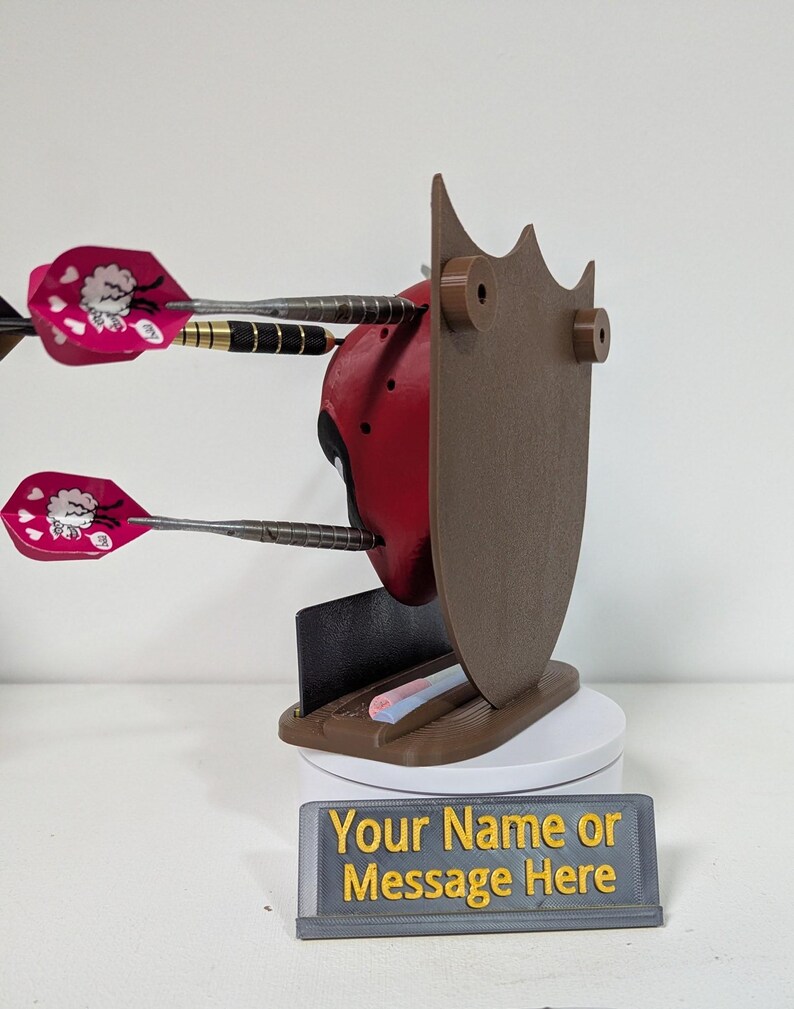 May include: A red and black Deadpool-themed dart holder with three darts. The holder has a brown shield-like back and a base with a slot for darts. A sign at the base reads "Your Name or Message Here."