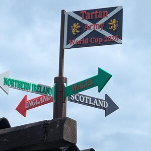 May include: A brown pole features a rectangular sign at the top with a dark blue background, a white saltire, two gold lions, and the text Tartan Army World Cup 2026. Below are four directional arrows labelled Northern Ireland, Wales, England, and Scotland.