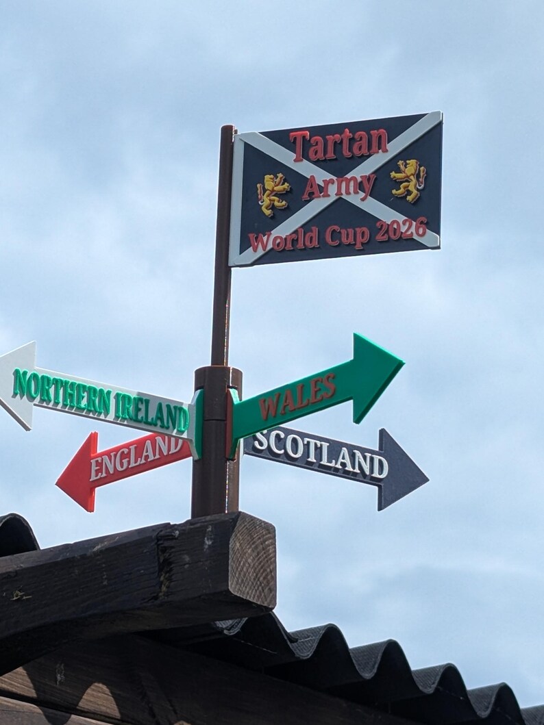 May include: A brown pole features a rectangular sign at the top with a dark blue background, a white saltire, two gold lions, and the text Tartan Army World Cup 2026. Below are four directional arrows labelled Northern Ireland, Wales, England, and Scotland.