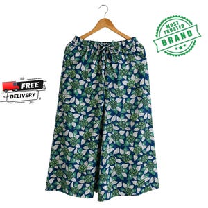 May include: Wide-leg pants featuring a dark blue background with a repeating pattern of green roses and white leaves. The pants have an elastic waistband with a drawstring tie and are displayed hanging on a wooden clothes hanger.