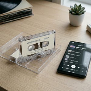 May include: A clear cassette tape with the text "YOUR MIX | VOL. II | SIDE A" and a smartphone displaying a music playlist titled "URBAN INDIE" are on a wooden surface. A small potted succulent is also visible.
