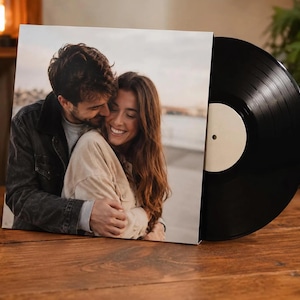 May include: A custom vinyl record featuring a square cardboard jacket with a photographic print of a smiling couple embracing. The record itself is black with a plain white center label, partially pulled out from the jacket to reveal the disc.