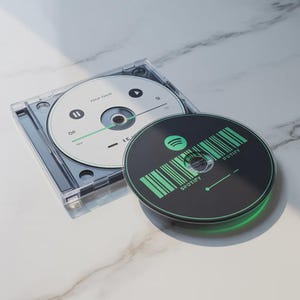 May include: A clear CD case with two CDs. One CD is white with a play button and a green line. The other CD is black with a green Spotify logo and barcode design. The CDs are on a white marble surface.