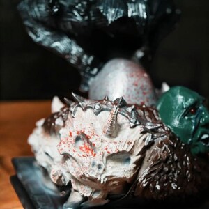 Puede incluir: Una figura detallada de una figura oscura de pie sobre una base con una calavera blanca y una cabeza de goblin verde. La calavera est&aacute; cubierta de pintura roja, dando la impresi&oacute;n de sangre.
