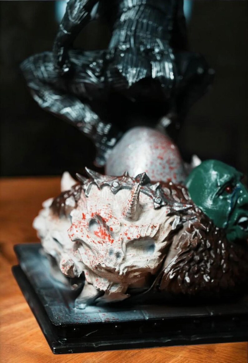 Puede incluir: Una figura detallada de una figura oscura de pie sobre una base con una calavera blanca y una cabeza de goblin verde. La calavera est&aacute; cubierta de pintura roja, dando la impresi&oacute;n de sangre.