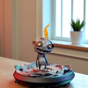 May include: A whimsical, gray figurine of a creature with a flame emanating from its head. The figure stands on a circular base with a colorful, textured design. The base has a black rim and is set on a wooden surface.