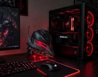 Puede incluir: Un casco de armadura Berserker negro y plateado con ranuras oculares rojas brillantes, colocado junto a una torre de computadora con iluminación interna roja, una tarjeta gráfica GeForce RTX y un monitor con una ilustración de fantasía.